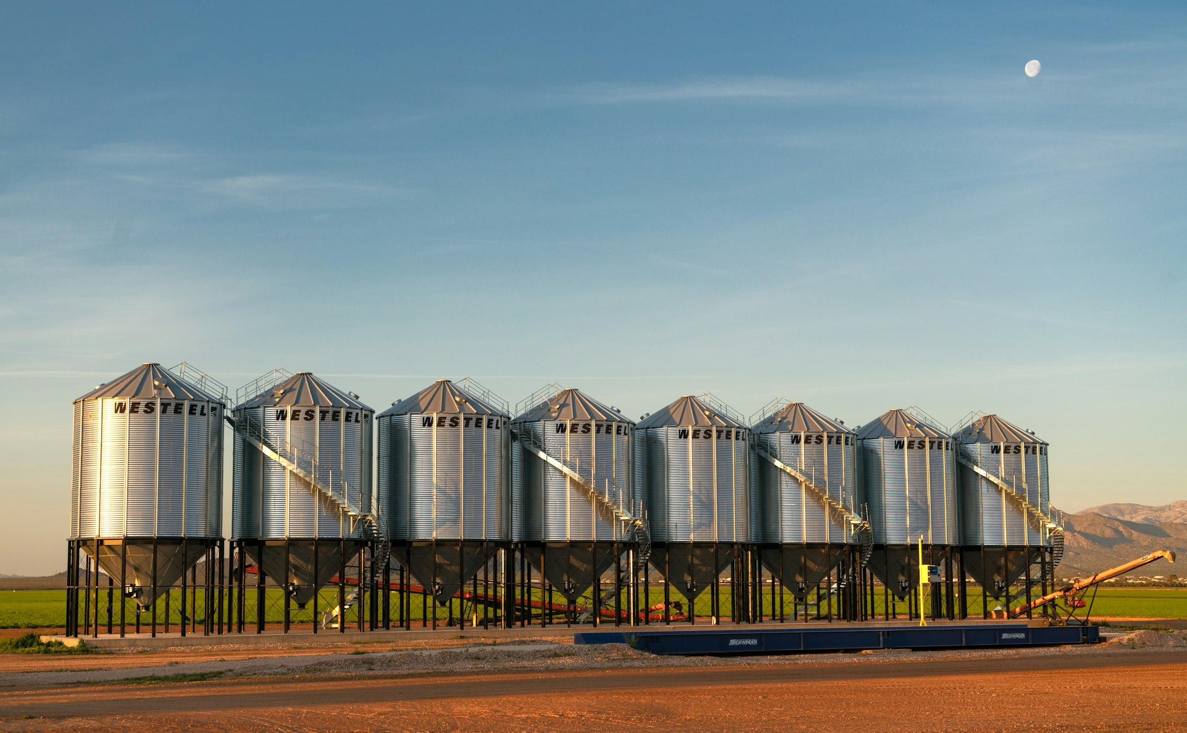A row of steel grain silos under a clear sky.